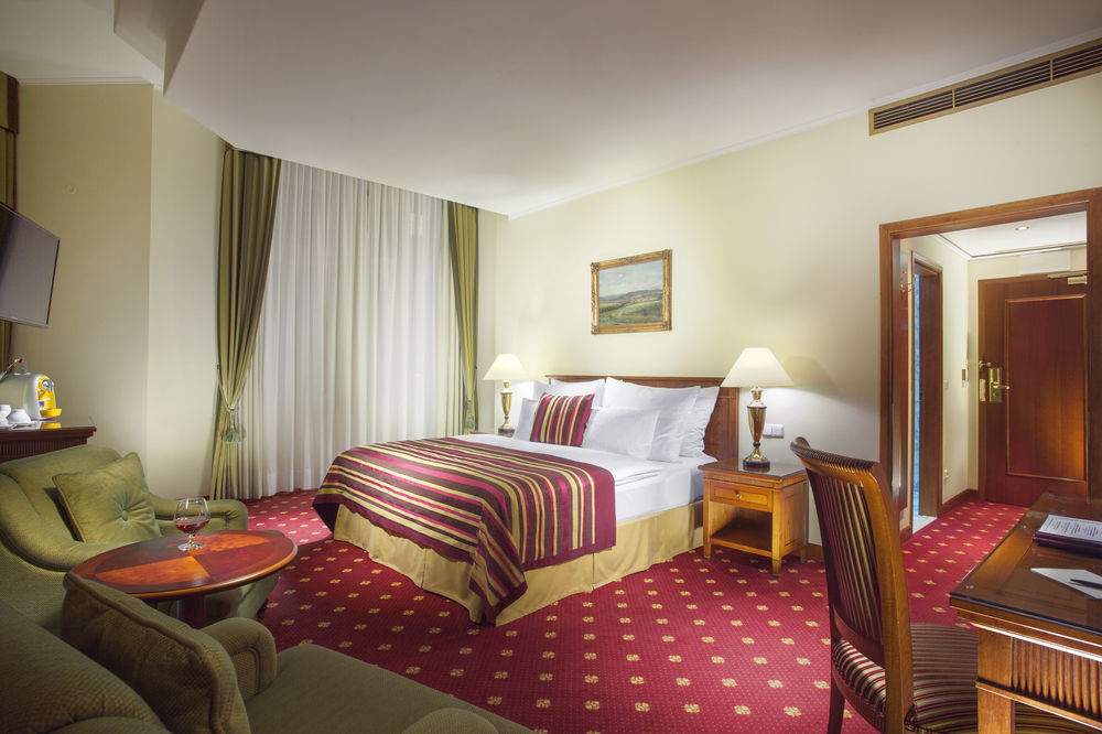 Art Nouveau Palace Hotel Praga Zewnętrze zdjęcie The photo shows a hotel room featuring a queen-sized bed adorned with striped bedding. There are two bedside lamps on either side of the bed. A cozy seating area includes a green couch and a small round table. The room has light-colored walls and a r